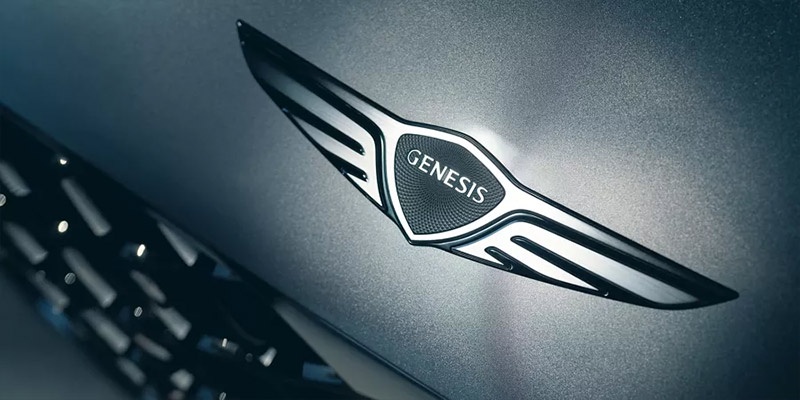 Close-up of the Genesis car logo with sleek silver wings on a dark background.
